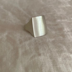 Pode incluir: Um anel de prata com uma parte superior de forma quadrada. A parte superior do anel tem um acabamento escovado.