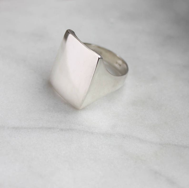 May include: A silver ring with a large, flat, square-shaped top. The ring is made of smooth, polished metal.