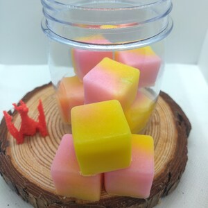 May include: A glass jar filled with pink and yellow wax melts. The wax melts are in the shape of cubes and are arranged in a pile. The jar is sitting on a wooden surface.