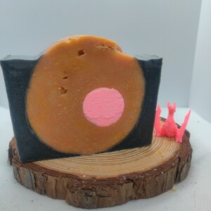 May include: A round bar of soap with a black and orange swirl design. The soap has a pink circle in the center. The soap is sitting on a wooden log slice.