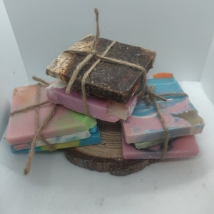 Puede incluir: Una pila de cinco barras de jabón hechas a mano atadas con cuerda. Las barras son de varios colores, incluyendo marrón, rosa, azul y verde. Los jabones están dispuestos sobre una rodaja de madera.