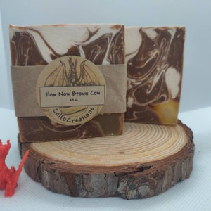 Puede incluir: Dos barras de jabón hechas a mano con un diseño marmoleado marrón y blanco. El jabón está etiquetado como "How Now Brown Cow" y "Zolfo Creations". El jabón está sentado sobre una rodaja de madera.