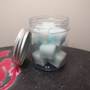 May include: A clear glass jar with a silver lid filled with small, white and blue wax melts. The jar is sitting on a black surface.