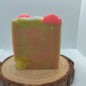 May include: A green and pink soap bar with a white swirl on top. The soap is sitting on a wooden round slice.