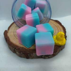 May include: Six pink and blue wax melts in the shape of cubes. The wax melts are in a clear glass jar on a wooden surface.