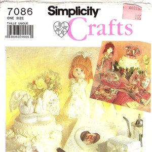 Könnte beinhalten: Simplicity Crafts Schnittmuster mit Hochzeits-Themen. Das Bild zeigt eine Puppe, einen verzierten Kuchen, Fotorahmen und eine kleine Box, alle in Weiß- und Cremetönen. Das Schnittmuster enthält den Text "7086" und "ONE SIZE".