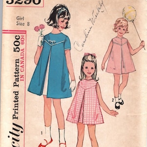 Peut inclure: Enveloppe de patron Simplicity vintage avec des illustrations de trois filles en robes. L'enveloppe affiche le numéro 5290, le texte "Girl Size 8" et le prix "50c". Les illustrations montrent des robes bleues, roses et à carreaux roses et blancs.