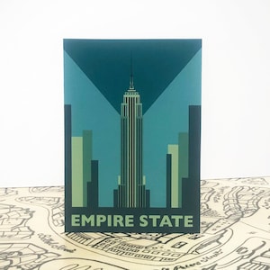 May include: A rectangular print featuring the Empire State Building in a teal and green color scheme, with the words "EMPIRE STATE" at the bottom. The print is set against a white background and rests on a map.