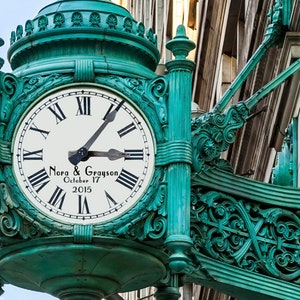 Op de afbeelding: Een turquoise groene klok met Romeinse cijfers en een wit gezicht. De wijzerplaat van de klok heeft de tekst "Nora & Grayson October 17 2015". De klok is gemonteerd op een decoratieve, sierlijke, turquoise groene beugel.