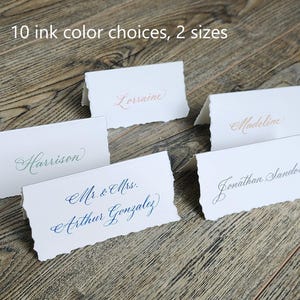May include: Five place cards with different names written in calligraphy. The place cards are on a wooden surface. The text on the place cards reads: "Lorraine", "Harrison", "Madeline", "Mr. & Mrs. Arthur Gonzalez", and "Jonathan Sandora".