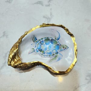 May include: A white oyster shell with a gold rim, painted with a blue and green sea turtle. The shell is a decorative dish or trinket tray.