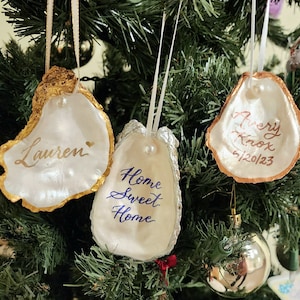 May include: Three white oyster shell ornaments with gold and copper accents hanging on a Christmas tree. The ornaments are personalized with the names "Lauren", "Avery Knox 6/20/2023", and "Home Sweet Home".