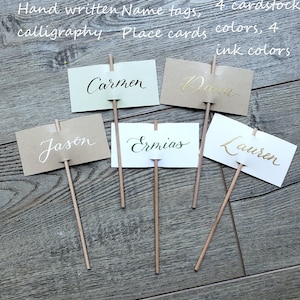 May include: Five place cards with names written in calligraphy on them. The cards are attached to wooden sticks and are arranged on a wooden surface. The names on the cards are Carmen, Dara, Jason, Ermias, and Lauren.