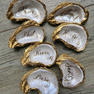 May include: Seven gold-painted oyster shells with white interiors. Each shell has a name written in gold calligraphy. The names are:  Shree K, John S, Phil B, Kevin M, Kevin M, Scott M, and Piero M.