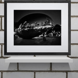 May include: Black and white framed photograph of a large, reflective, bean-shaped sculpture. The sculpture reflects a cityscape at night, with lights and buildings visible. The photograph is displayed on a white shelf against a brick wall.