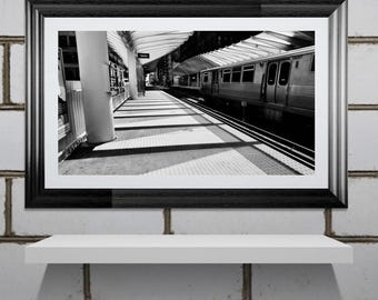 Könnte beinhalten: Schwarz-weiß gerahmte Fotografie eines U-Bahnhofs. Das Bild zeigt einen Zug auf den Gleisen, einen Bahnsteig und architektonische Details. Die Fotografie wird auf einem weißen Regal vor einer weißen Backsteinmauer ausgestellt.