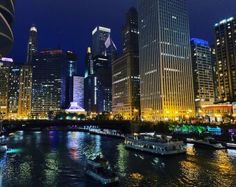 May include: A night view of the Chicago skyline with tall buildings lit up with lights. The Chicago River is in the foreground with several boats on the water.