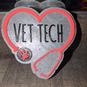 May include: A gray heart-shaped emblem with the words "VET TECH" in black. A red stethoscope design frames the heart, and a paw print is visible. The emblem is likely a badge or decoration.