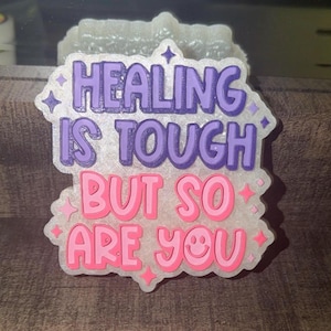 May include: A white, glittery, semi-translucent plaque featuring the purple text HEALING IS TOUGH and pink text BUT SO ARE YOU. The design includes small star and smiley face accents, with a raised, layered appearance and a scalloped edge border.
