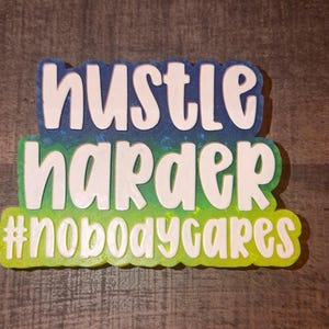 May include: A decorative sign featuring the white text "hustle harder #nobodycares" in a rounded, bold font. The background consists of a gradient that transitions from a deep blue at the top to a bright lime green at the bottom with a glittery finish.