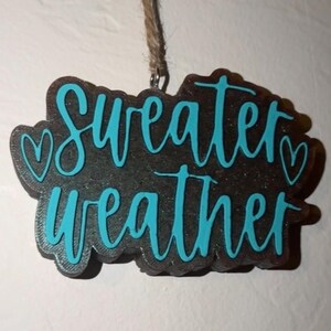 May include: A decorative sign with the words "Sweater Weather" in teal script. The sign is dark brown with a textured appearance and two teal hearts. It hangs from a natural twine string.