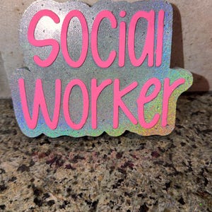 May include: A silver-colored, glittery sign with the words "SOCIAL WORKER" in bright pink, outlined in pink. The sign has a holographic, iridescent border. The background is a speckled, granite-like surface.
