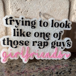 May include: A white and pink ornament with the text "trying to look like one of those rap guys girlfriends" in black and pink lettering. The ornament has a small silver loop at the top for hanging.