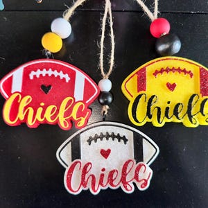 May include: Three red, yellow, and white football-shaped ornaments with the word "Chiefs" written in black. Each ornament has a small heart in the center.