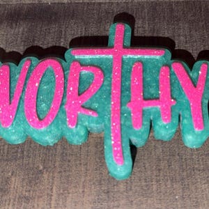 May include: A teal-colored, glittery decorative piece with the word "WORTHY" in bright pink, and a pink cross. The letters and cross are outlined in a darker teal. The piece is set against a wooden background.