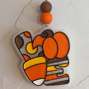 May include: A fall-themed hanging decoration featuring a pumpkin and candy corn design. The decoration is made of clear acrylic with orange, yellow, brown, and white accents. It is suspended by a natural twine string and wooden beads.