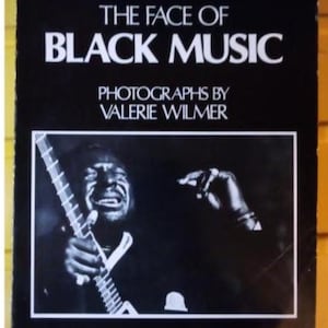 Könnte beinhalten: Schwarzes Buchcover mit dem Titel "THE FACE OF BLACK MUSIC" in weißen Großbuchstaben. Es zeigt eine Schwarz-Weiß-Fotografie eines Musikers mit einem Instrument. Fotografien von Valerie Wilmer, Einführung von Archie Shepp.