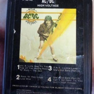 May include: An AC/DC "High Voltage" 8-track tape. The cover features a band member playing a guitar with a yellow lightning bolt graphic. Song titles include "It's A Long Way To The Top" and "T.N.T."