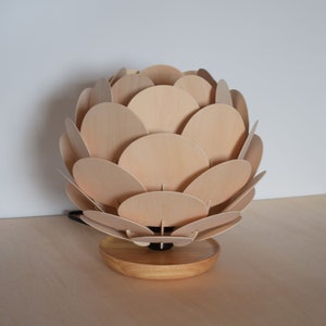 Puede incluir: Una l&aacute;mpara de mesa de madera con un dise&ntilde;o &uacute;nico que se asemeja a una pi&ntilde;a. La pantalla de la l&aacute;mpara est&aacute; hecha de m&uacute;ltiples c&iacute;rculos de madera superpuestos, creando un aspecto texturizado y org&aacute;nico. La base es un simple disco de madera.