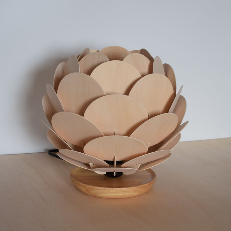 Puede incluir: Una l&aacute;mpara de mesa de madera con un dise&ntilde;o &uacute;nico que se asemeja a una pi&ntilde;a. La pantalla de la l&aacute;mpara est&aacute; hecha de m&uacute;ltiples c&iacute;rculos de madera superpuestos, creando un aspecto texturizado y org&aacute;nico. La base es un simple disco de madera.