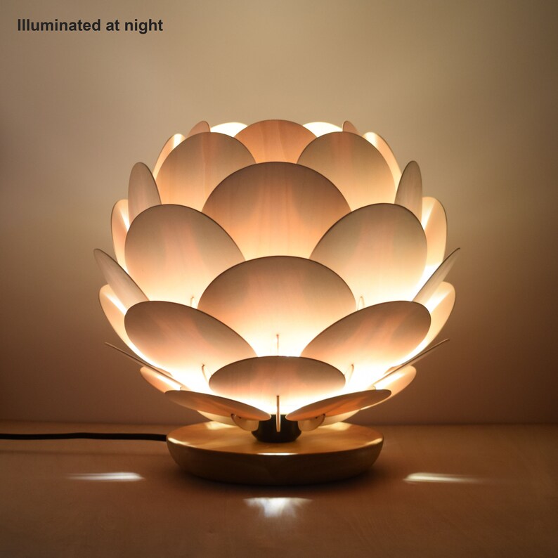 Puede incluir: Una l&aacute;mpara de mesa de madera con un dise&ntilde;o &uacute;nico que se asemeja a una flor. La l&aacute;mpara est&aacute; hecha de m&uacute;ltiples piezas de madera superpuestas, creando un efecto de capas. La l&aacute;mpara est&aacute; iluminada, proyectando un brillo c&aacute;lido.