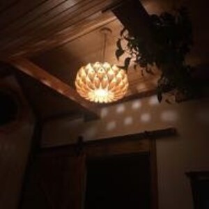 May include: A wooden pendant light fixture with a unique, geometric design. The light is hanging from a ceiling with wood paneling and casts a warm glow.