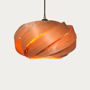 Może przedstawiać: Nowoczesna lampa wisząca o unikalnym designie. Lampa posiada drewniany klosz złożony z warstwowych, zakrzywionych pasków jasnobrązowego drewna. Wnętrze świeci ciepłym pomarańczowym światłem i wisi na brązowym kablu.