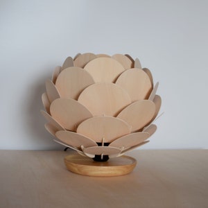 Puede incluir: Una l&aacute;mpara de mesa de madera con un dise&ntilde;o &uacute;nico que se asemeja a un capullo de flor. La pantalla de la l&aacute;mpara est&aacute; hecha de m&uacute;ltiples c&iacute;rculos de madera superpuestos, creando un efecto de capas. La base es un simple disco de madera.