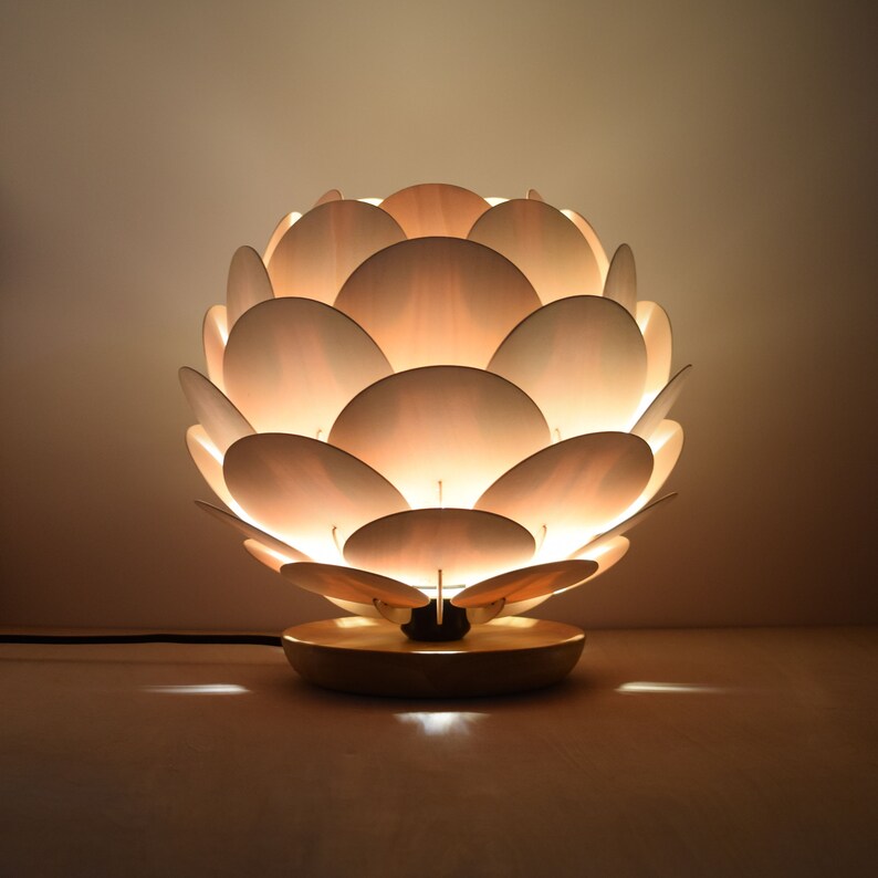 Puede incluir: Una l&aacute;mpara de mesa blanca con un dise&ntilde;o &uacute;nico que se asemeja a un capullo de flor. La pantalla de la l&aacute;mpara est&aacute; hecha de m&uacute;ltiples p&eacute;talos blancos superpuestos, creando un brillo suave y c&aacute;lido.