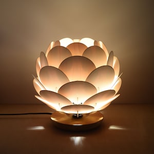 Puede incluir: Una l&aacute;mpara de mesa de madera blanca con un dise&ntilde;o &uacute;nico que se asemeja a una flor. La l&aacute;mpara est&aacute; encendida y proyecta un brillo c&aacute;lido.