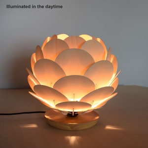 Puede incluir: Una l&aacute;mpara de mesa de madera con un dise&ntilde;o &uacute;nico que se asemeja a un capullo de flor. La l&aacute;mpara est&aacute; hecha de m&uacute;ltiples piezas de madera delgadas y redondeadas que crean un efecto de capas. La l&aacute;mpara est&aacute; iluminada, proyectando un c&aacute;lido resplandor.