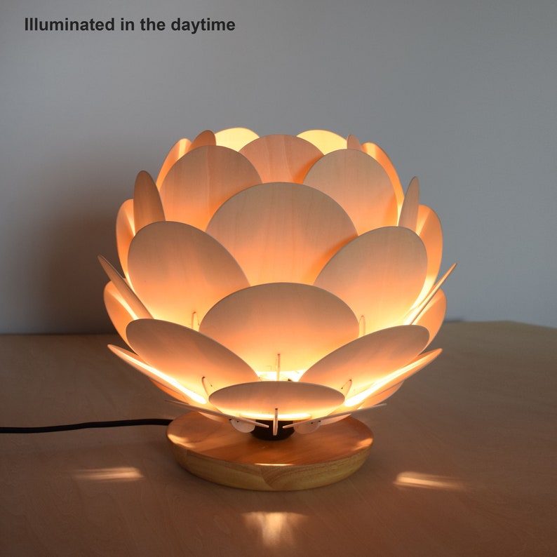 Puede incluir: Una l&aacute;mpara de mesa de madera con un dise&ntilde;o &uacute;nico que se asemeja a un capullo de flor. La l&aacute;mpara est&aacute; hecha de m&uacute;ltiples piezas de madera delgadas y redondeadas que crean un efecto de capas. La l&aacute;mpara est&aacute; iluminada, proyectando un c&aacute;lido resplandor.