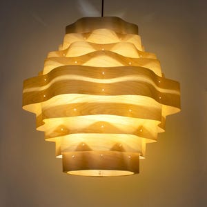 May include: A pendant light fixture constructed from layered, wavy bands of light-colored wood veneer. The structure features a series of horizontal, undulating tiers that create a tiered, organic shape, with small pinpoints of light visible between the layers.