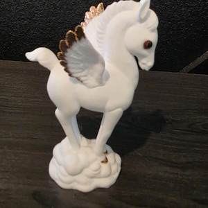 May include: A white ceramic Pegasus figurine with brown eyes, gold accents on the mane, and brown-tipped wings. The mythical creature stands on a cloud-shaped base, showcasing a detailed and elegant design. This decorative item is ideal for home decor.