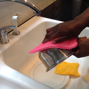 May include: A person is washing a metal bowl with a pink sponge in a white kitchen sink. A yellow sponge is on a white plate in the sink.