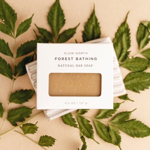 May include: A natural bar soap, labeled "Slow North Forest Bathing," is displayed in a white box with a window. The soap is light brown and rests on a white, ridged soap dish. Green leaves surround the soap, and the box indicates the soap weighs 4.5 oz / 127 g.