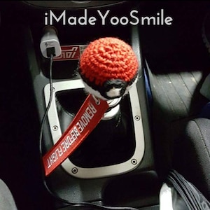 May include: A red and white crocheted shift knob with a black band, resembling a Pokeball. A red tag with white text that reads "REMOVE BEFORE FLIGHT" hangs below the knob. The text "iMade YooSmile" is at the top.