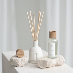 Puede incluir: Un difusor de cerámica blanca con palitos de madera y una botella de vidrio transparente con una tapa de madera y el texto "HOME SCENTS CLOUDS" sobre una superficie blanca texturizada.