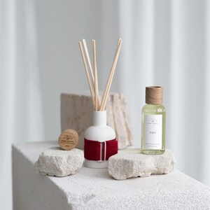 Peut inclure: Un diffuseur en céramique blanche avec un ruban rouge et des bâtonnets en bois, un flacon en verre transparent avec un bouchon en bois et le texte "JOY" sur l'étiquette, et un bouchon de bouteille en bois avec le texte "HOME SCENTS" dessus. Les articles sont sur une surface texturée blanche.