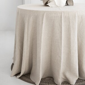 May include: A round, beige linen tablecloth draped over a table with a white ceramic bowl filled with dried greenery in the center.